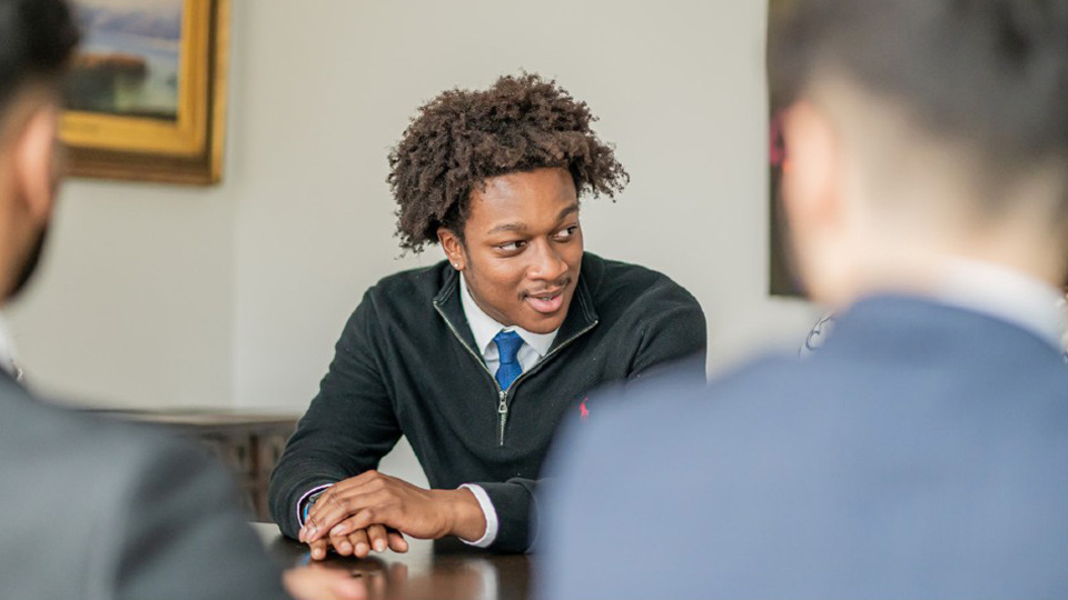 A person wearing a dark sweater and blue tie, sits at a table.